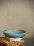 May include: A small ceramic bowl featuring a glossy blue interior and rim that transitions into a raw, unglazed beige base. The bowl has a smooth, rounded shape with a distinct color separation between the glazed upper portion and the matte lower section.