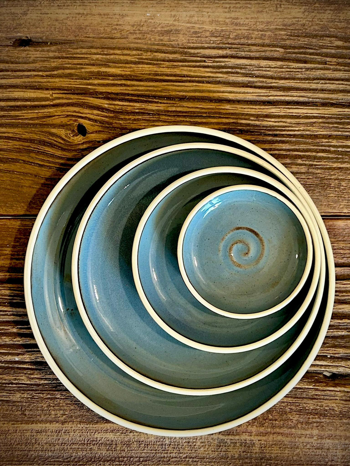 May include: A set of four nested ceramic plates and a small bowl, each featuring a smooth, dusty blue glaze with subtle speckles and a distinct cream-colored rim. The smallest bowl in the center displays a hand-painted brown spiral design.