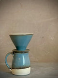 May include: A ceramic coffee mug with a matching conical pour-over dripper stacked on top. The set features a glossy blue glaze on the upper sections, transitioning into a dark brown band, and a solid off-white base on the mug.