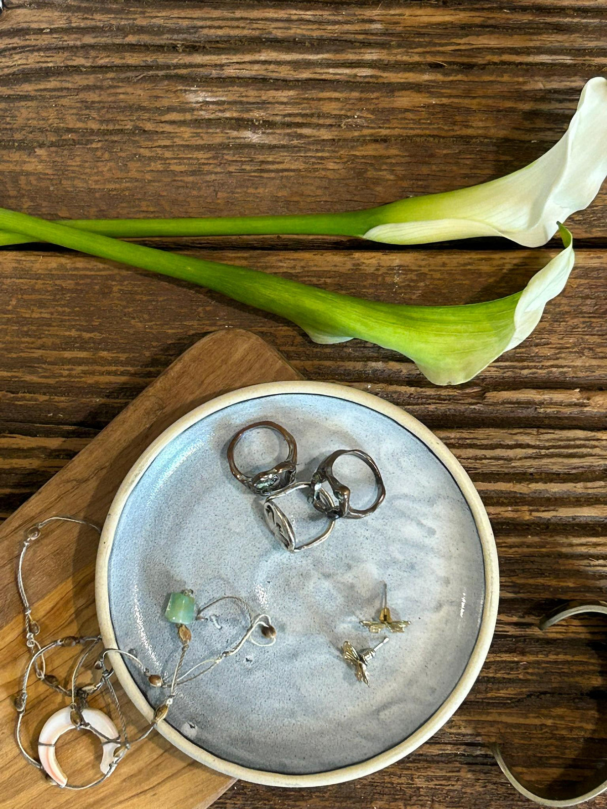May include: A round, light blue ceramic dish containing three dark metal rings, a pair of small metal stud earrings, and a delicate silver-toned chain necklace with a white crescent moon pendant and a small green bead accent.