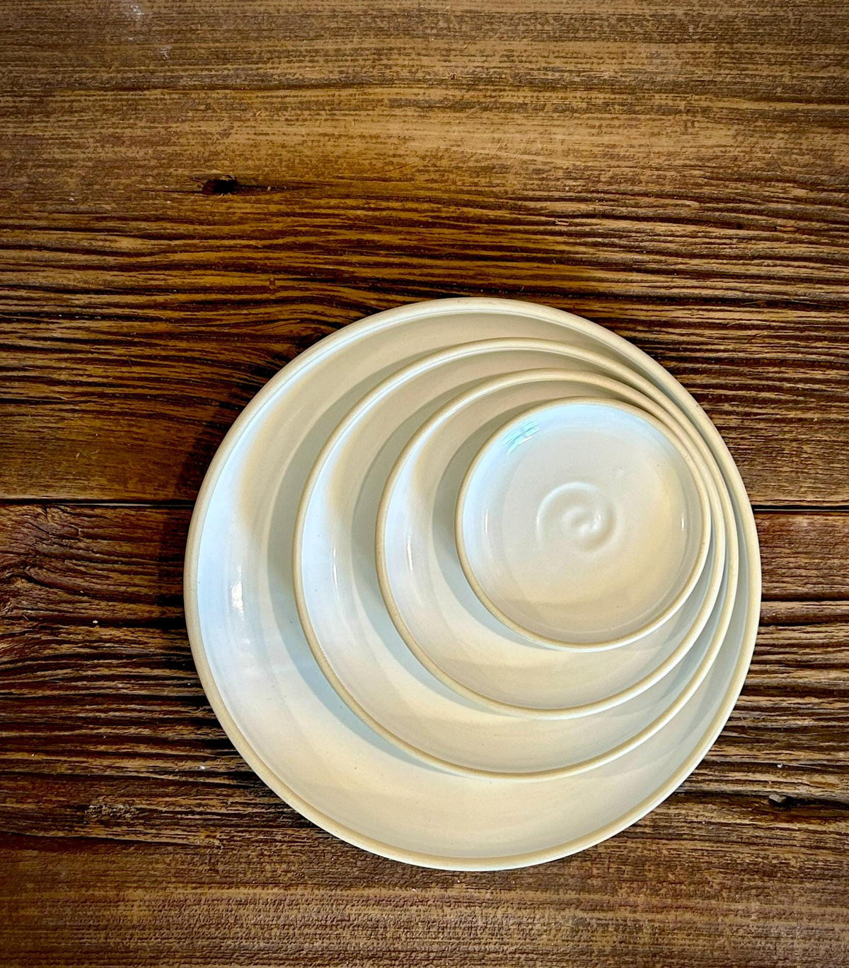 May include: A set of four nested circular ceramic plates in a glossy off-white finish. The plates are stacked from largest to smallest, with the smallest plate featuring a subtle spiral indentation in the center and a raised rim on each piece.