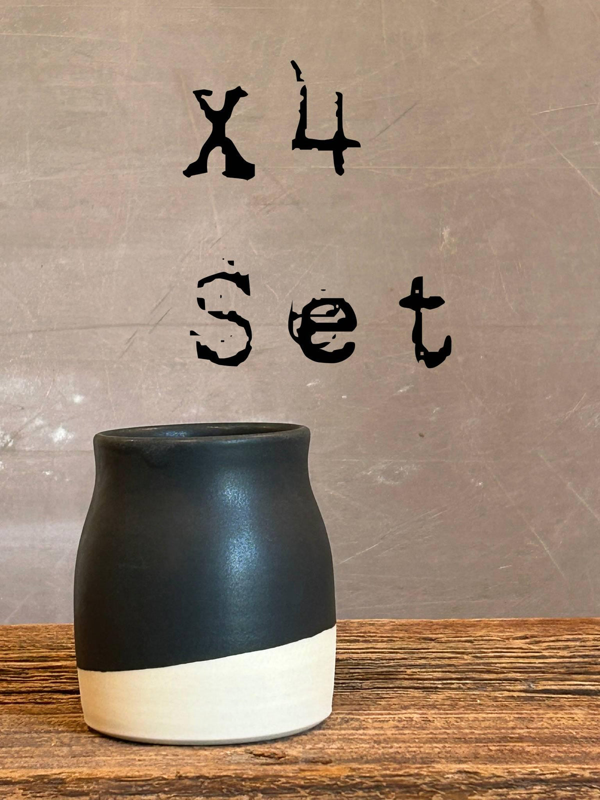 May include: A set of four ceramic vessels featuring a two-tone design with a matte black upper section and a contrasting off-white base. The text "x4 Set" is displayed in black lettering above the vessel, which has a rounded body and a wide opening.