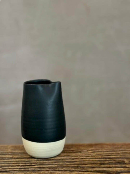 May include: A ceramic pitcher featuring a matte black upper section and a contrasting off-white base. The vessel has a cylindrical body with a slightly tapered neck and a small, integrated pouring spout at the rim, showcasing a two-tone design.