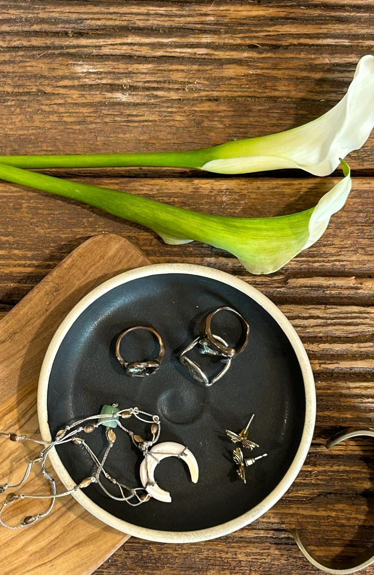 May include: A round black ceramic dish with a white rim containing two metal rings, a pair of gold-toned butterfly stud earrings, and a delicate necklace featuring a white crescent moon pendant and small metallic beads arranged in a circular pattern.