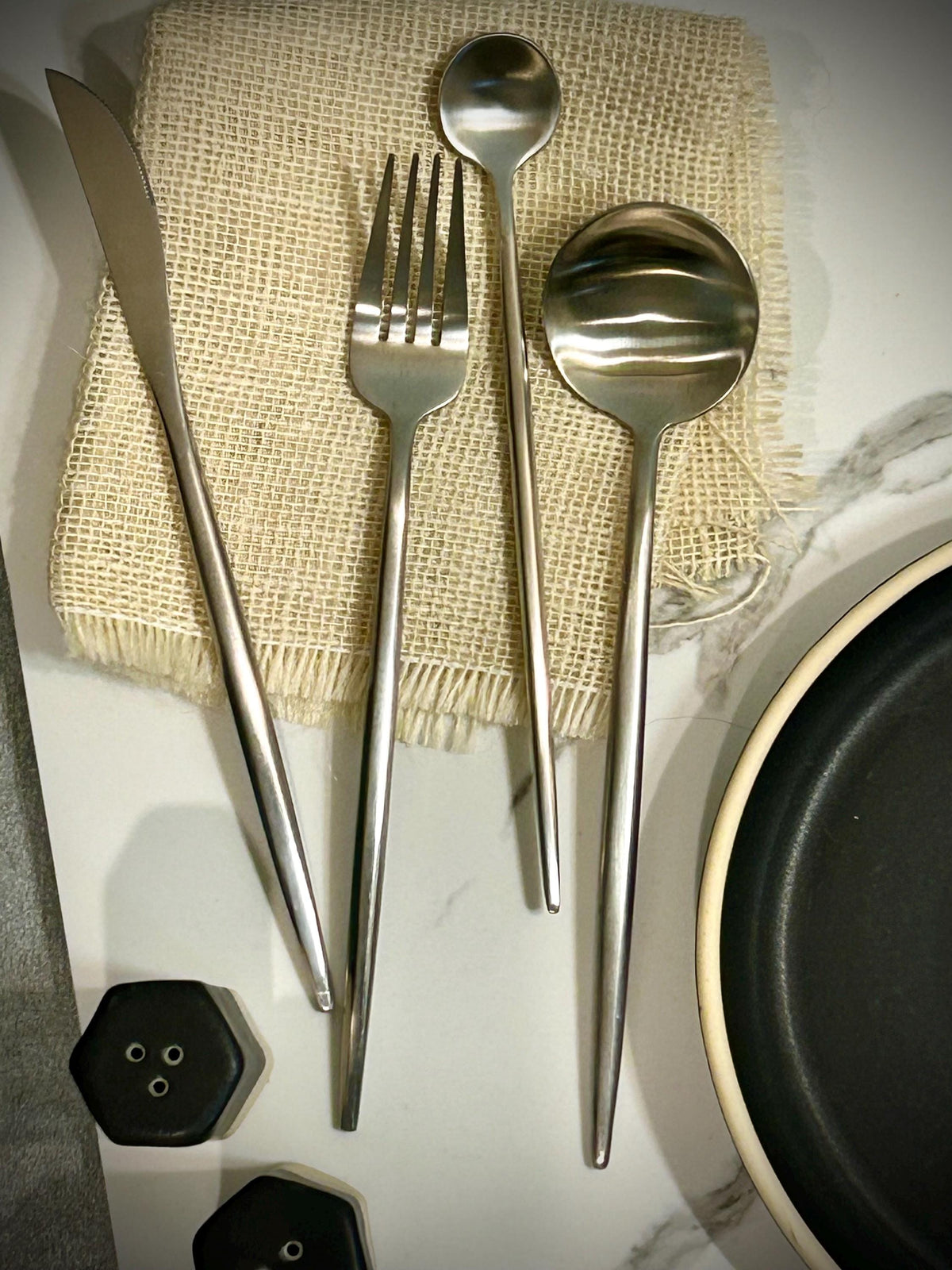 May include: A set of four stainless steel utensils including a dinner knife, a dinner fork, a small round spoon, and a large round spoon. The utensils feature long, slender, tapered handles with a smooth, matte metallic finish and clean lines.