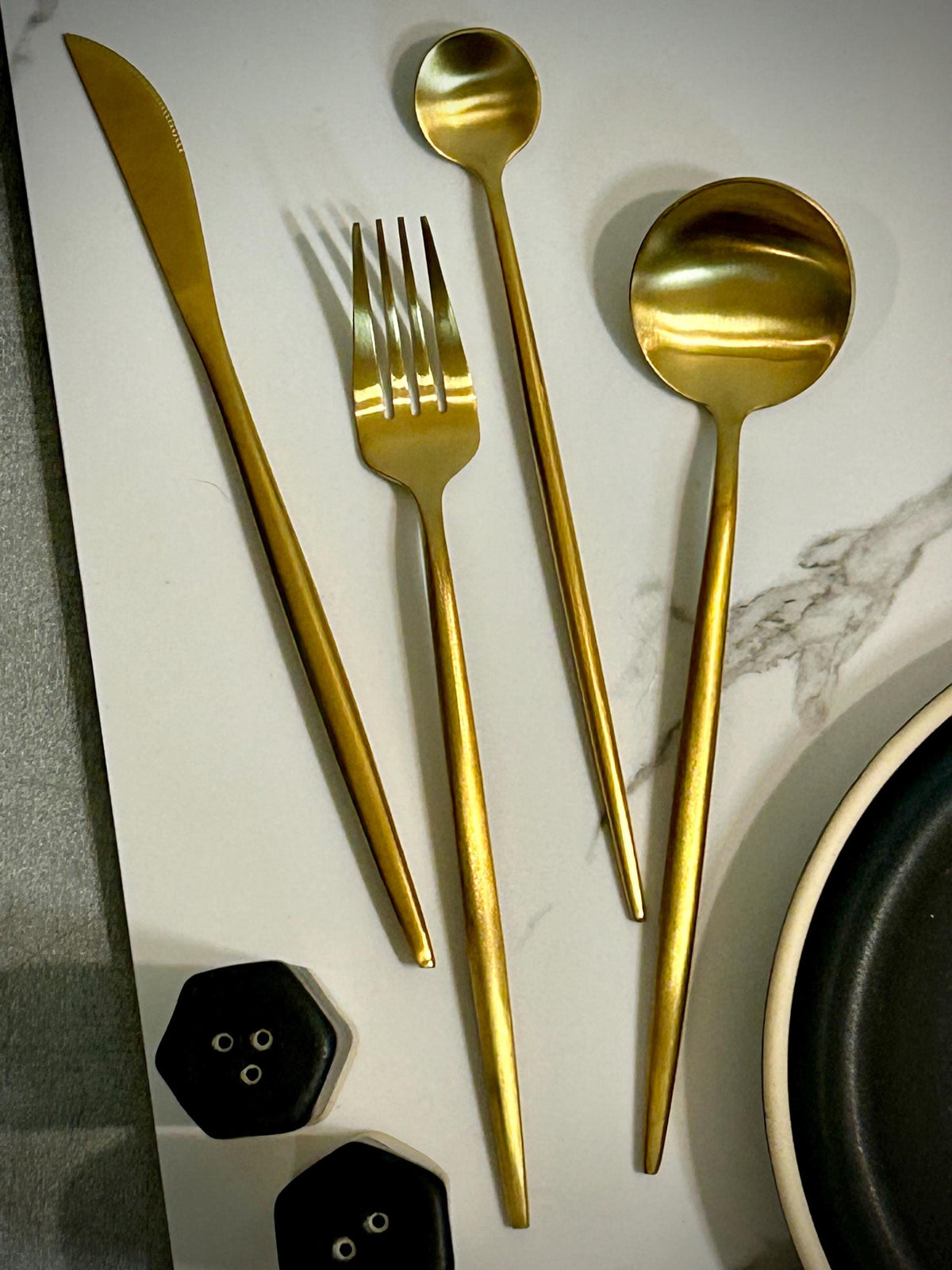 May include: A set of four gold-colored stainless steel utensils including a dinner knife, a dinner fork, a small round spoon, and a large round spoon. The utensils feature long, slender handles and a smooth, matte metallic finish.